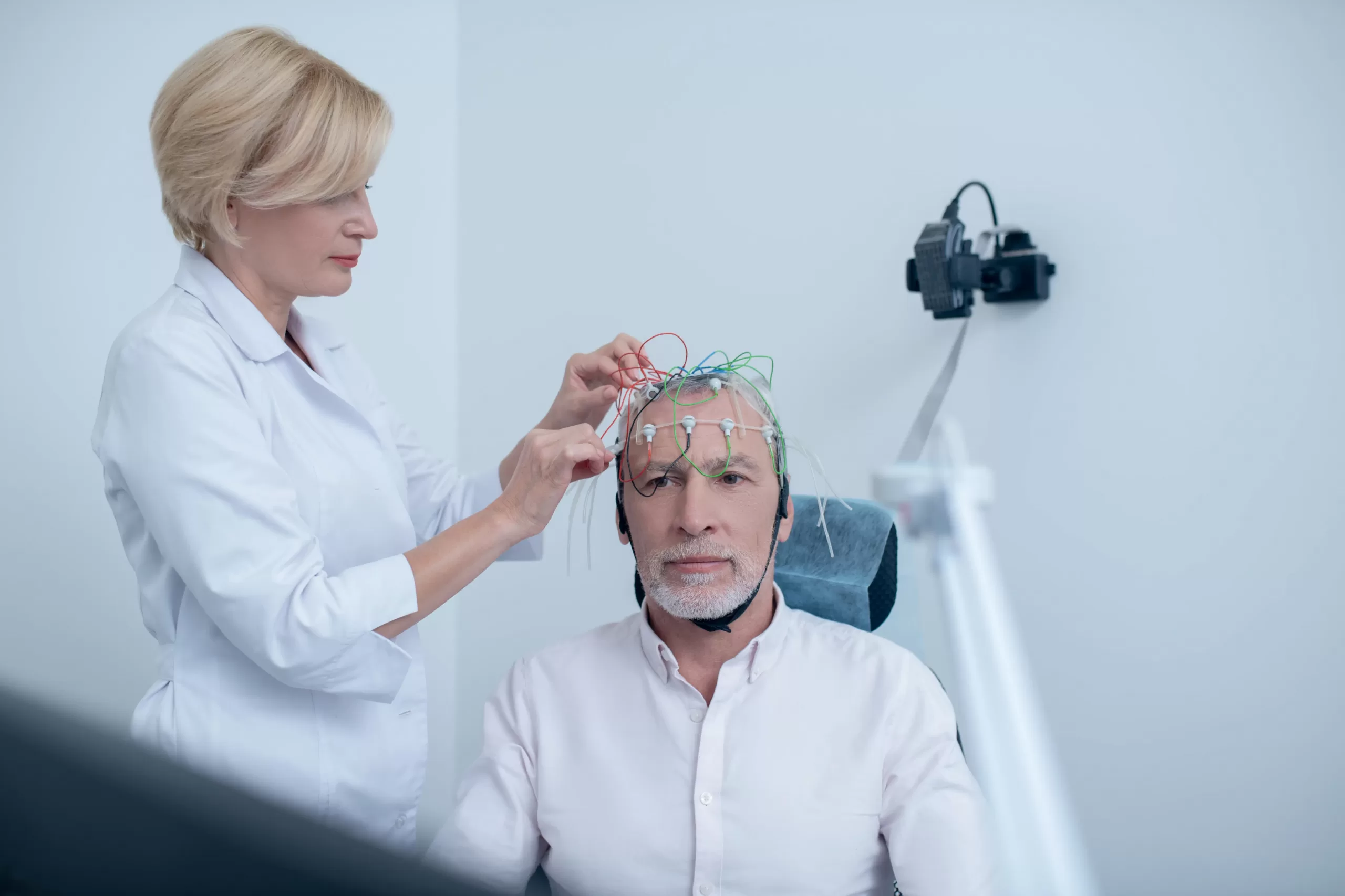 Female neurologist adjusting electrodes on gray-haired male patient head undergoing electroencephalogram